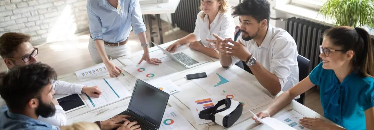 Team meeting in a modern office, with diverse professionals discussing data and strategies around a table filled with charts and graphs. A woman leads the presentation, while others engage with laptops and a virtual reality headset. Natural light floods the room, enhancing the collaborative atmosphere.